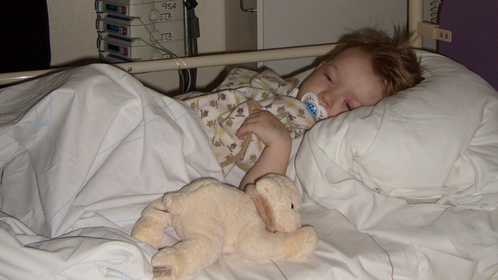 Noel ligger i en sjukhuss&auml;ng, med napp i munnen, snuttefilt och godsedjur. Foto.